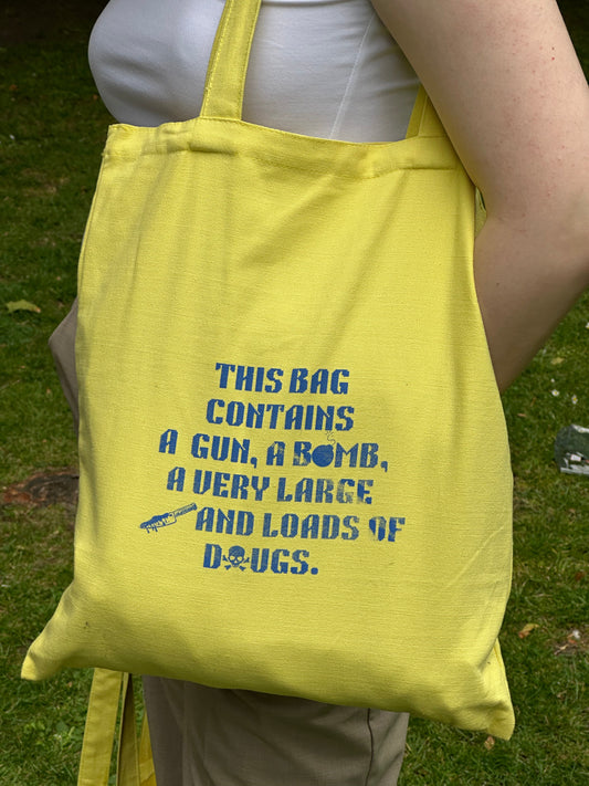 Yellow tote bag with blue hazard-themed stencil text and icons, designed by Tantrm